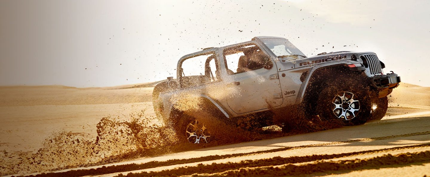 A 2020 Jeep Wrangler Rubicon with its top off kicking up sand as it drives over a hill.