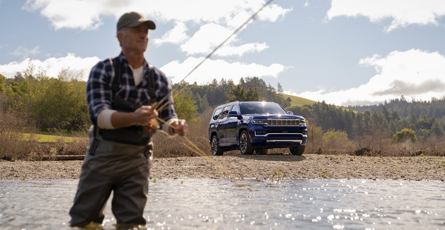 The 2022 Grand Wagoneer parked on a gravel riverbank with a fisherman in the foreground.